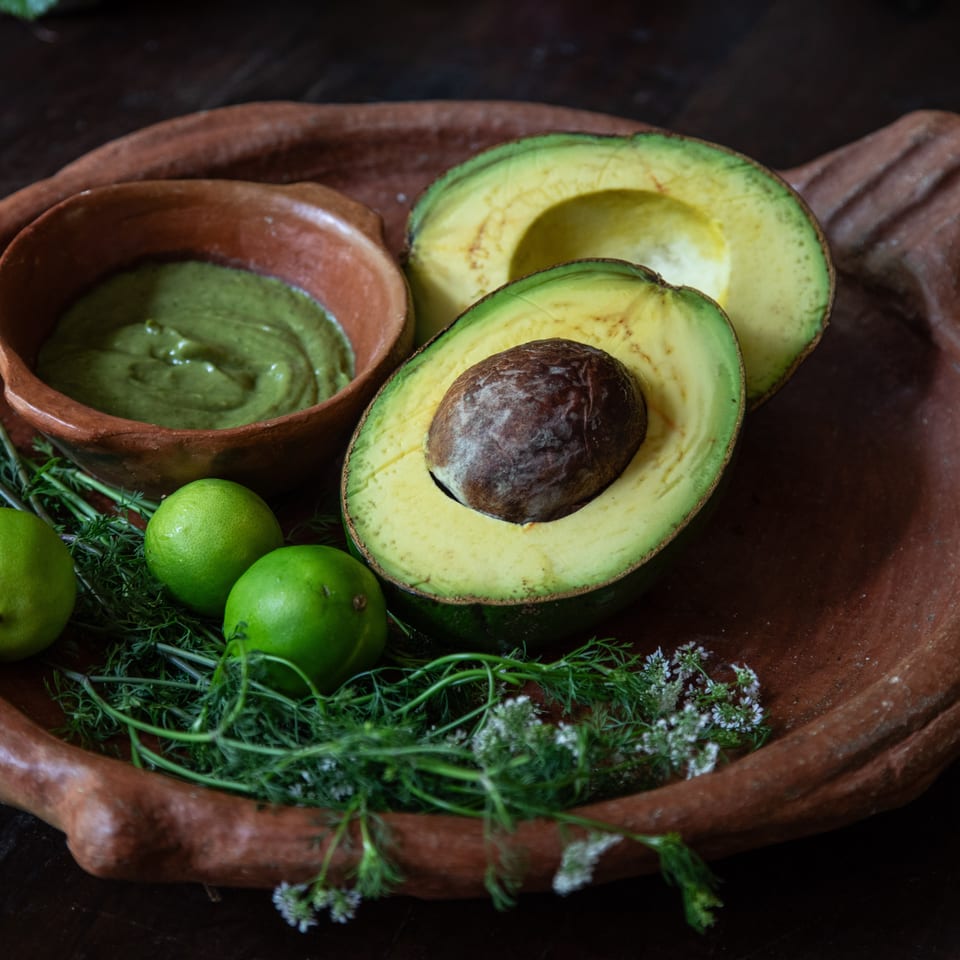 Halved avocados, limes, herbs, and a bowl of green sauce arranged on a rustic clay platter.