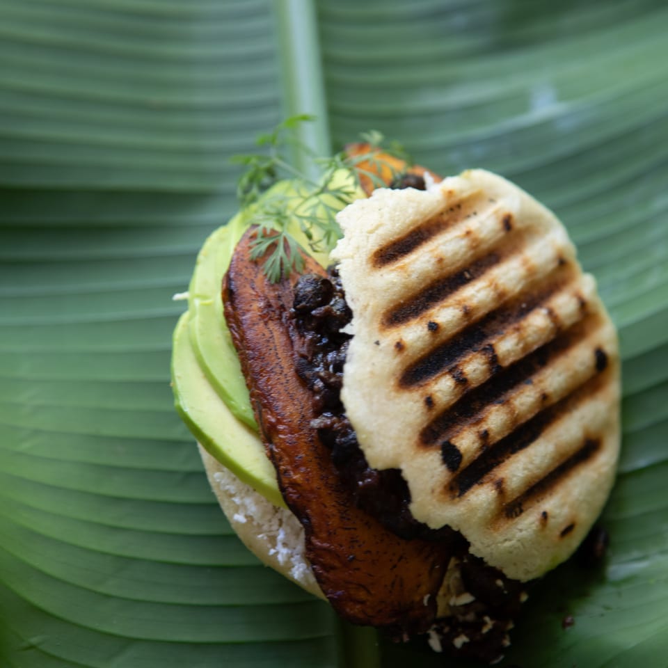 Close-up of a grilled sandwich filled with avocado, roasted vegetables, black beans, and fresh herbs on a banana leaf.