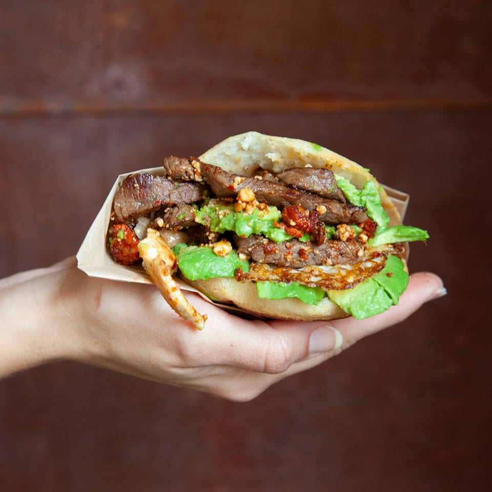 Hand holding a loaded sandwich with grilled beef, avocado, crispy egg, vegetables, and chili sauce in a paper tray.