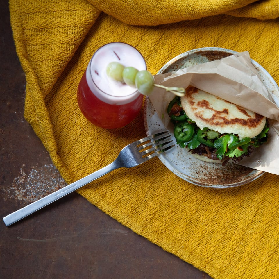 Overhead view of a stuffed arepa with cilantro and jalapeños, a red grape-garnished drink, and fork on a mustard-yellow cloth.