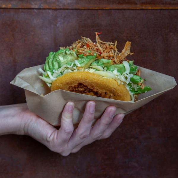 Hand-held toasted taco filled with guacamole, shredded cheese, crispy vegetables, and red peppers in a paper wrapper.